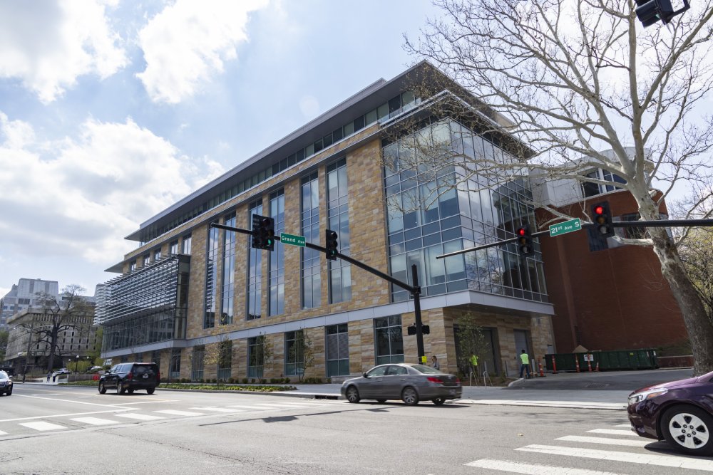 Street view of a large modern building with glass windows and stone accents, located at the corner of Grand Avenue and 21st Avenue. Several cars are stopped at a red light in front of the building, and a few pedestrians can be seen near the entrance. The sky is partly cloudy.